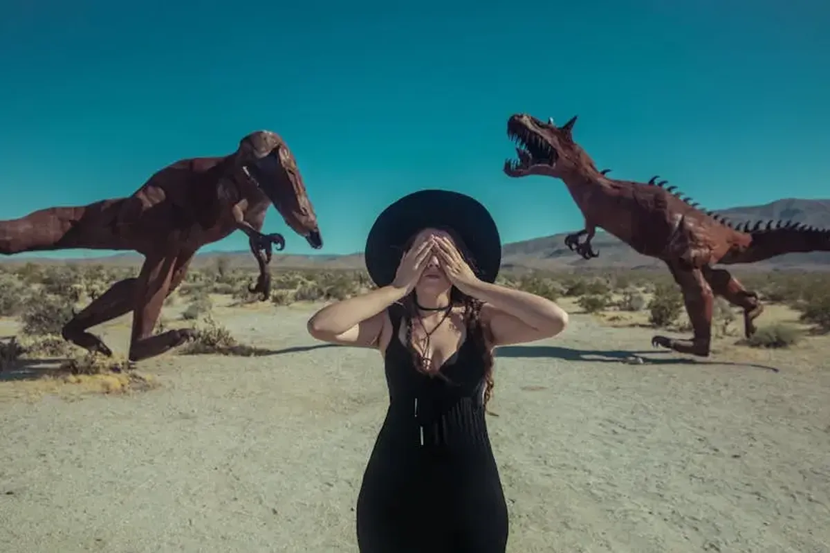 A woman covers her eyes among life-sized dinosaur sculptures in a desert setting.