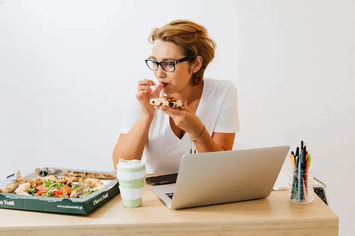A woman at a laptop eating pizza.